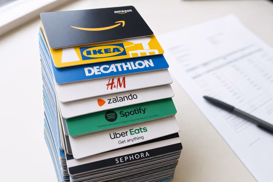 Una pila alta de tarjetas de regalo sobre un escritorio junto a una factura, con Amazon, IKEA, Decathlon, H&M, Zalando, Spotify, Uber Eats y Sephora legibles