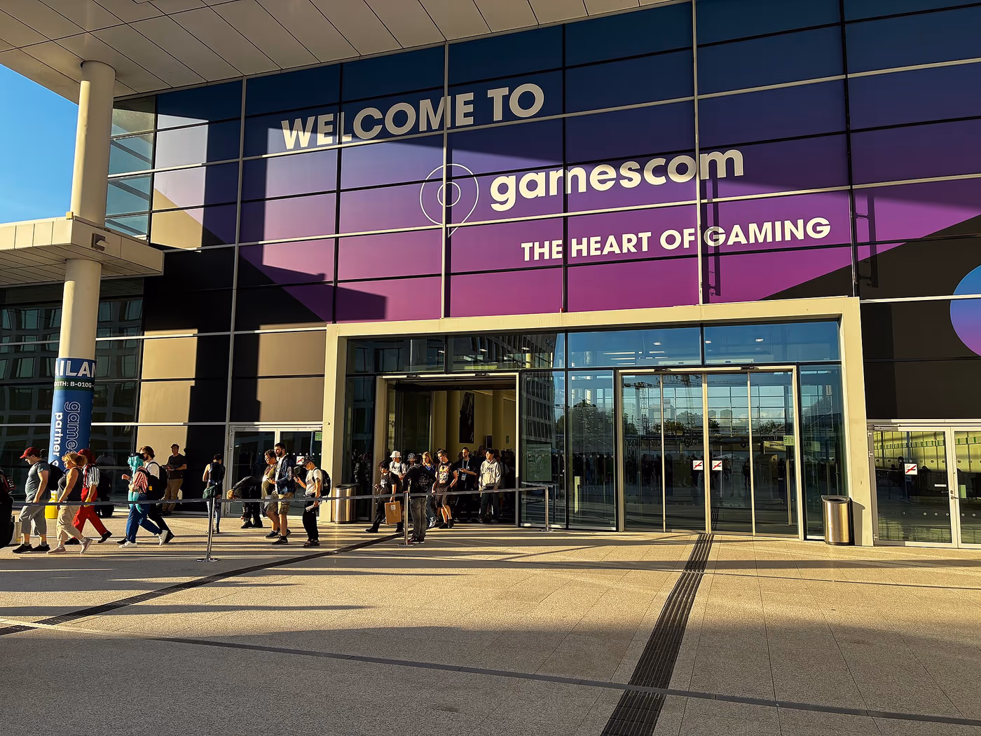 The Koelnmesse main entrance at gamescom 2025 in evening light, wrapped in the purple "Welcome to gamescom - the heart of gaming" facade, with visitors queuing outside