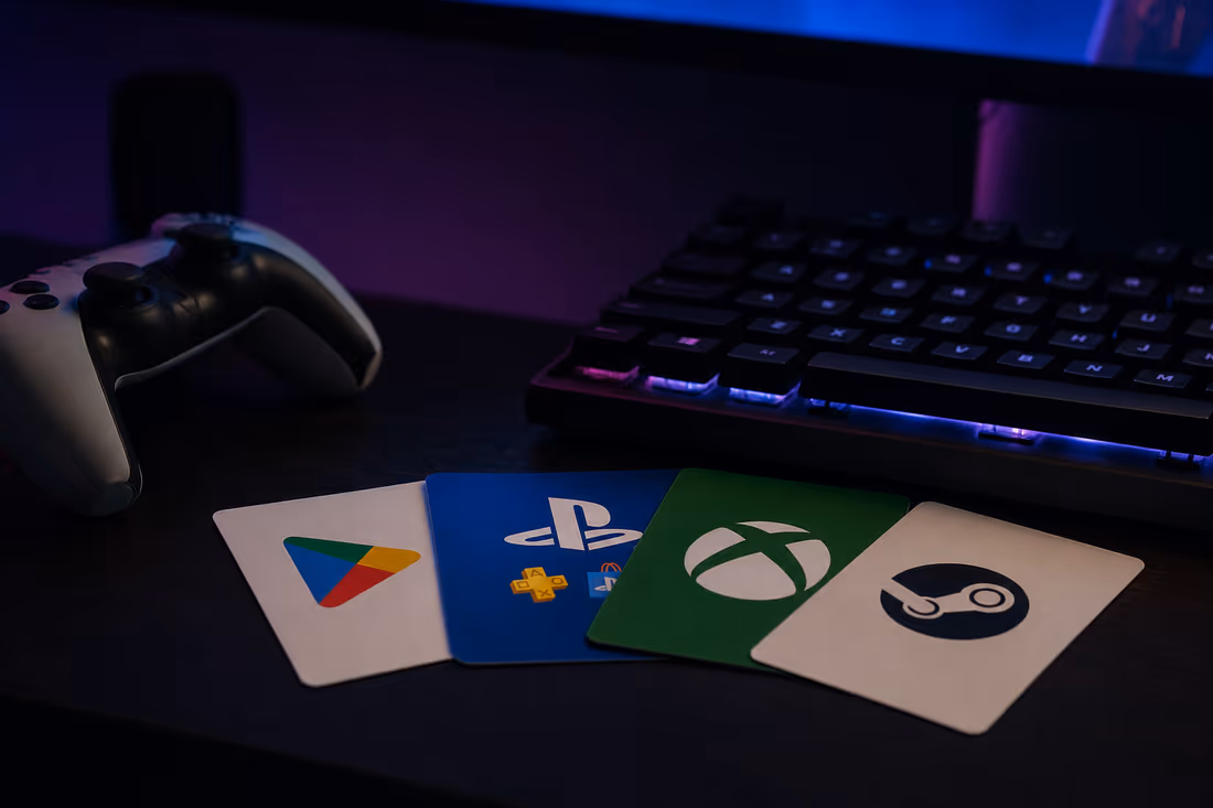 A gaming desk lit at night with a backlit keyboard, a controller, and Steam, Xbox, PlayStation and Google Play gift cards fanned out
